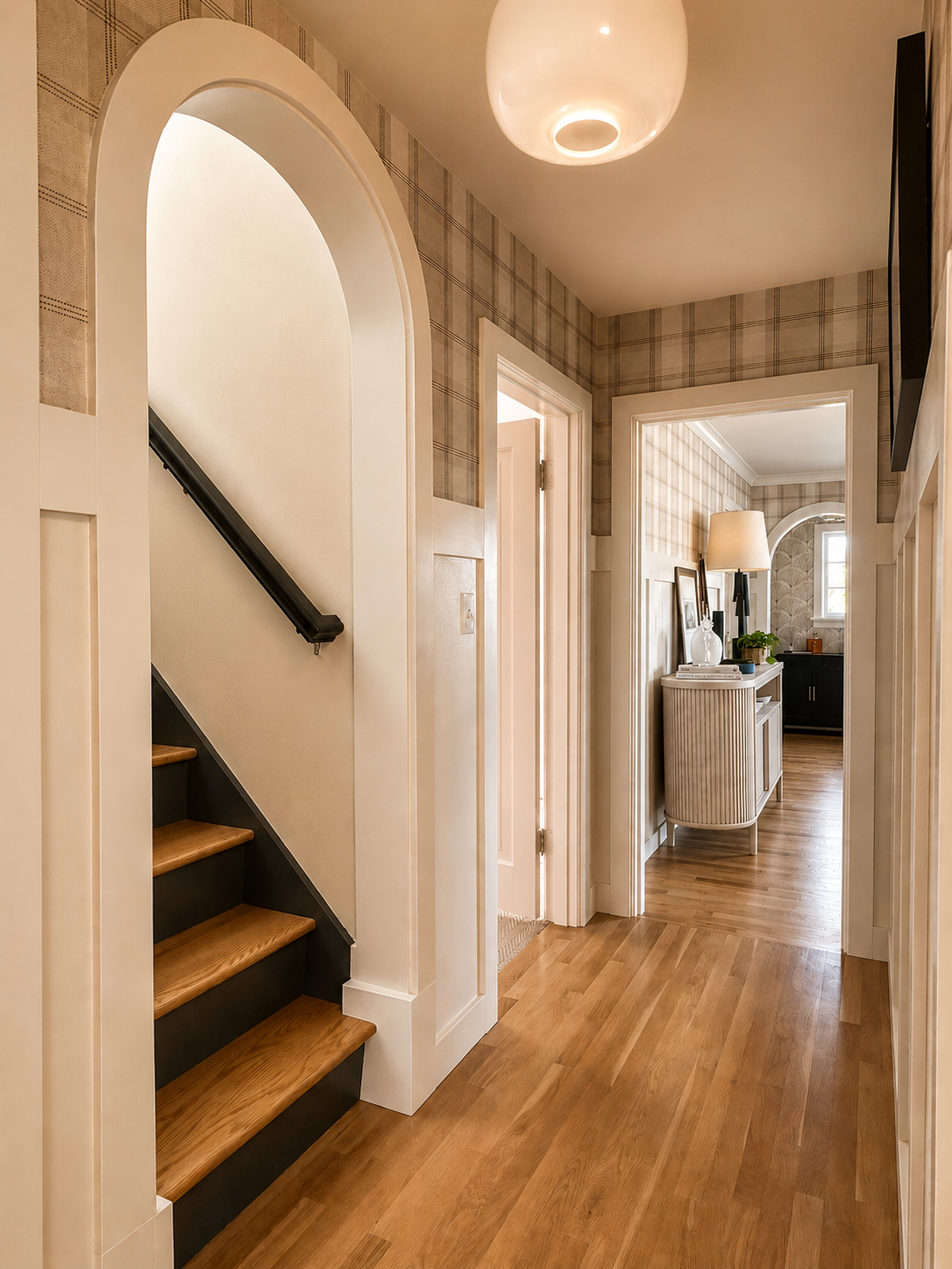 Beck Road hallway — arched stair opening, plaid wallpaper, warm oak floors, reeded console, globe pendant, view through to bar alcove