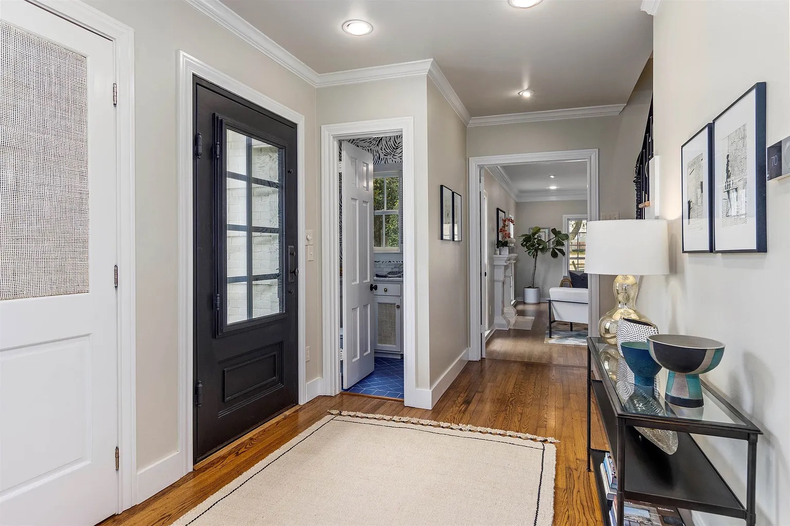 Tyler Estate entry hall — warm oak floors, black front door with glass, crown moulding, console table with art objects, view to living room