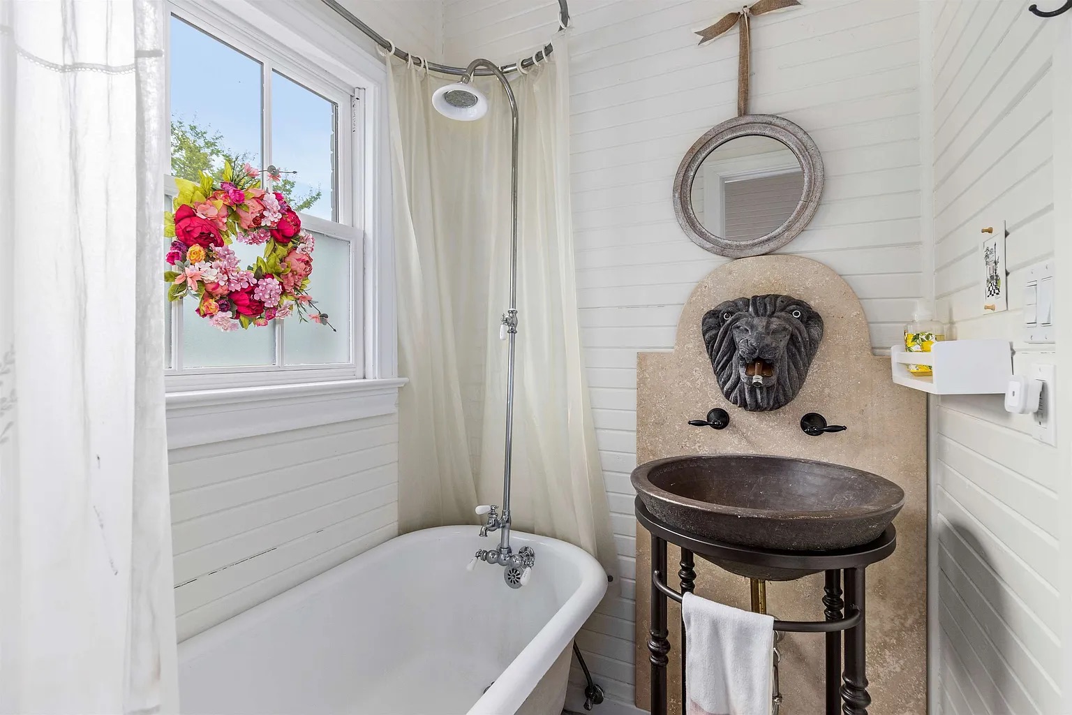 Tyler Estate guest bath — clawfoot tub with overhead shower, pink floral window wreath, stone lion-head vessel sink on iron stand, white shiplap walls