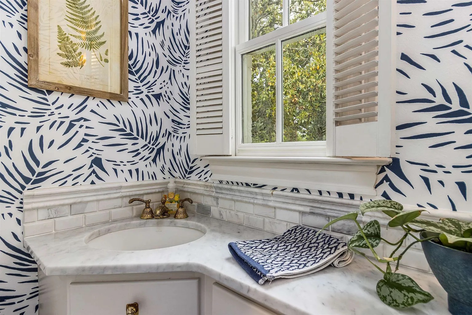 Tyler Estate powder bath — bold navy palm leaf wallpaper, marble vanity with brass fixtures, corner sink, shuttered window with garden view