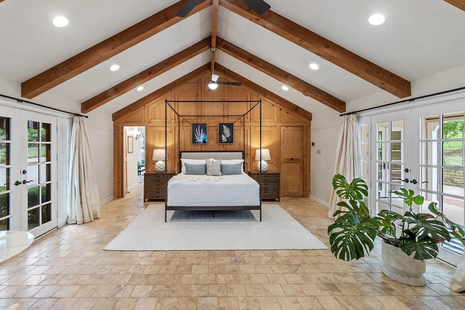 Tyler Estate primary bedroom — vaulted ceiling with exposed timber trusses, four-poster iron bed, warm cedar accent wall, French doors to grounds, monstera plant