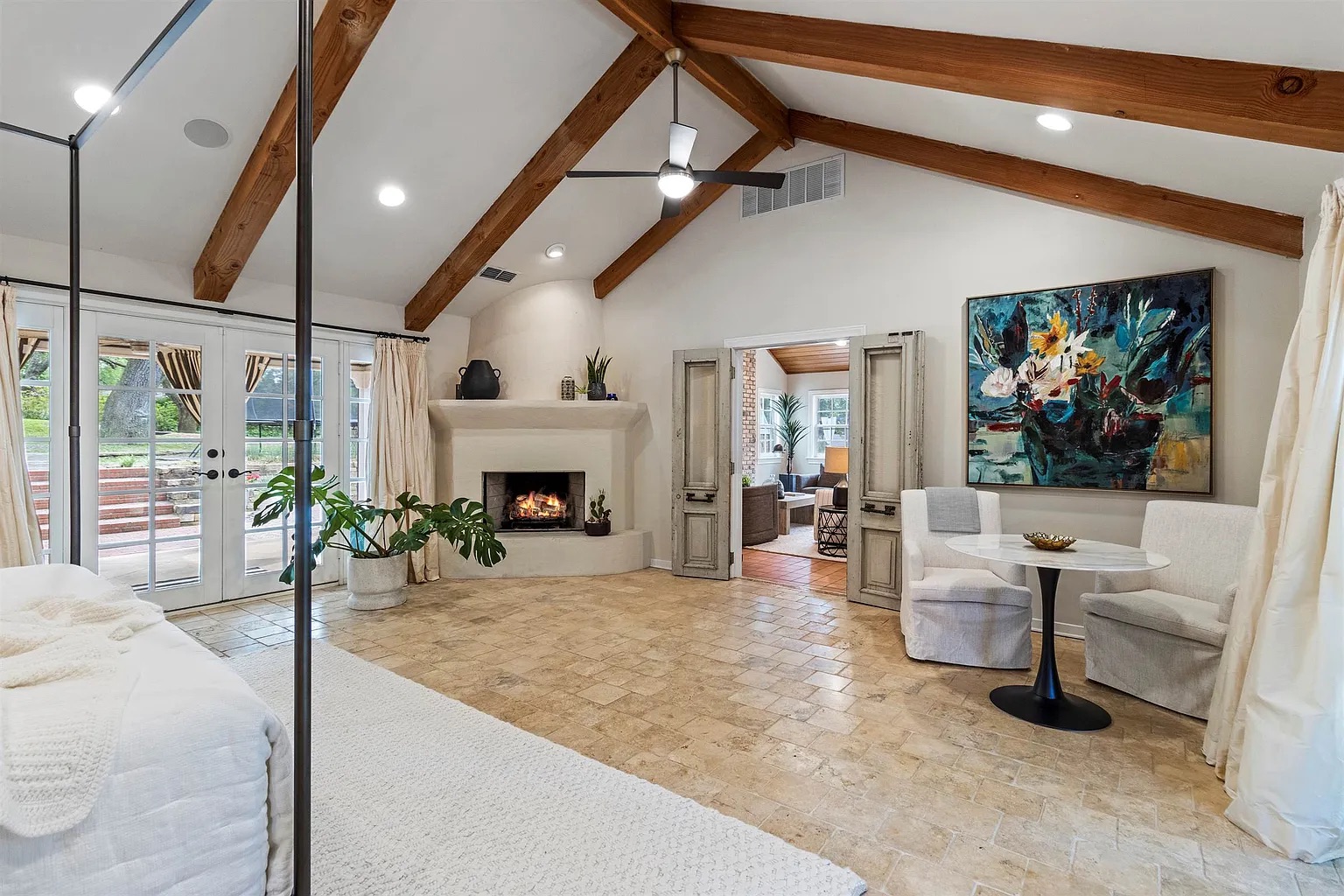 Tyler Estate primary suite fireplace — adobe-style corner fireplace with timber trusses, white slipcovered chairs, monstera plant, blue abstract painting