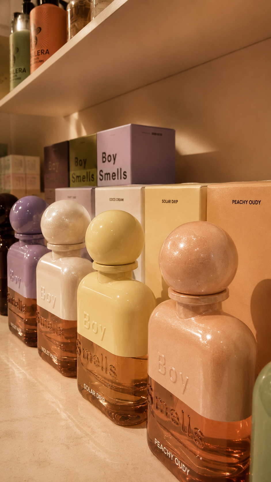 Lunamira Boy Smells fragrance display — sculptural glitter-finish perfume bottles in lavender, white, chartreuse, and peach on backlit marble-effect shelving